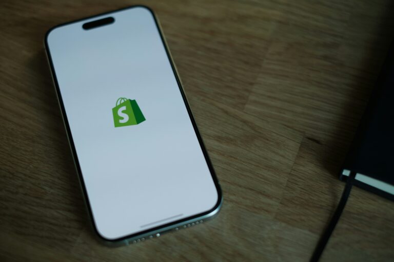 A cellphone sitting on a wooden table, with the Shopify logo visible against a white background on the device's screen.