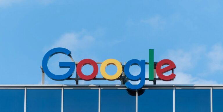 Google's distinctive wordmark logo is displayed above an office building, against a blue sky backdrop.